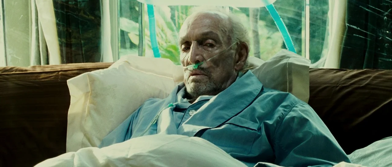 Slice (2018) movie still: medium — A man rests in a bed positioned before a large window with white curtains. He wears a blue pajama shirt and a…