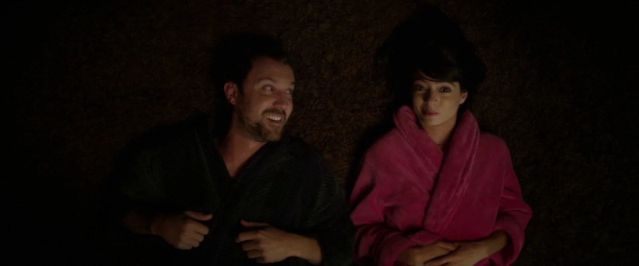 Seven Stages to Achieve Eternal Bliss (2018) movie still: medium — Two people lie on their backs on a textured brown shag carpet surface. They wear…