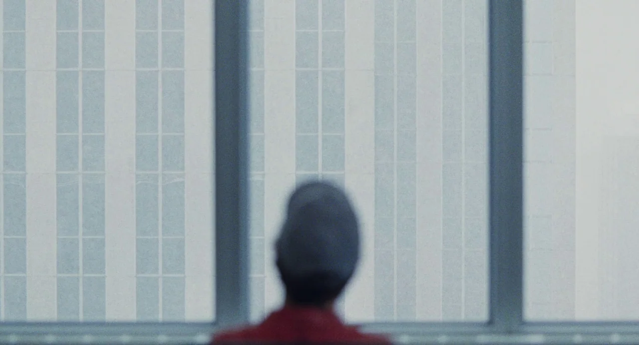 Dream Scenario (2023) movie still: medium — A view through a large glass window shows a modern building facade opposite. An individual wearing a…