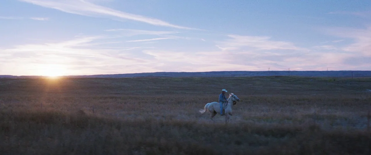 The Rider (2017) movie still: wide — A vast, open plain stretches toward a distant, low mountain range under a sunset sky. A lone cowboy on a light…
