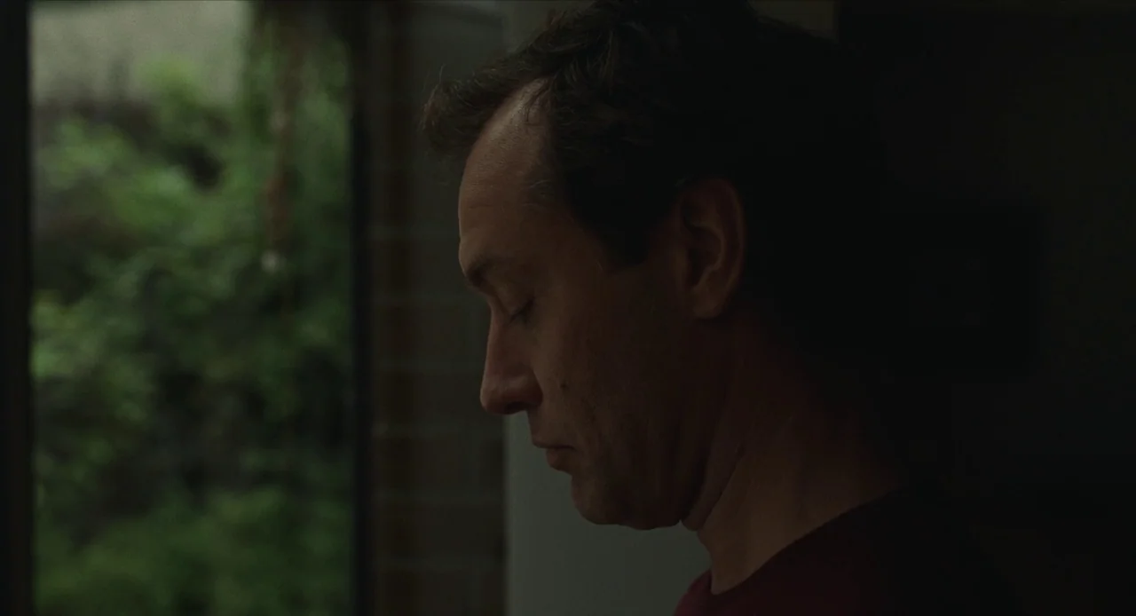 The Nest (2020) movie still: closeup — A man stands indoors next to a window overlooking a leafy green exterior. He faces left with his eyes closed,…