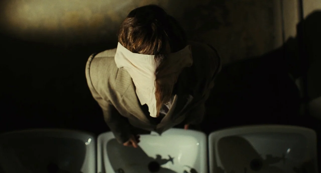 The Double (2013) movie still: medium — A dimly lit washroom contains a row of clean white porcelain sinks. A man wearing a tan blazer leans over a…