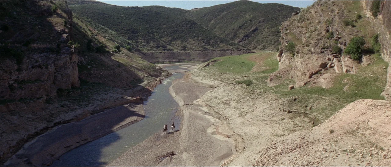 Texas, Adios (1966) movie still: wide — A rugged canyon features steep, rocky walls surrounding a winding river and riverbed. Two riders on…