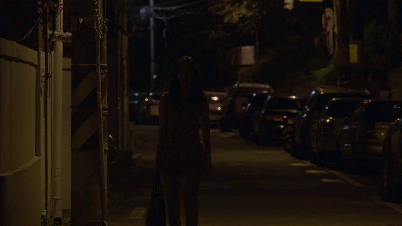 Yourself and Yours (2016) movie still: fullbody — A dimly lit narrow road at night is lined with parked vehicles and walls. A young woman walks…