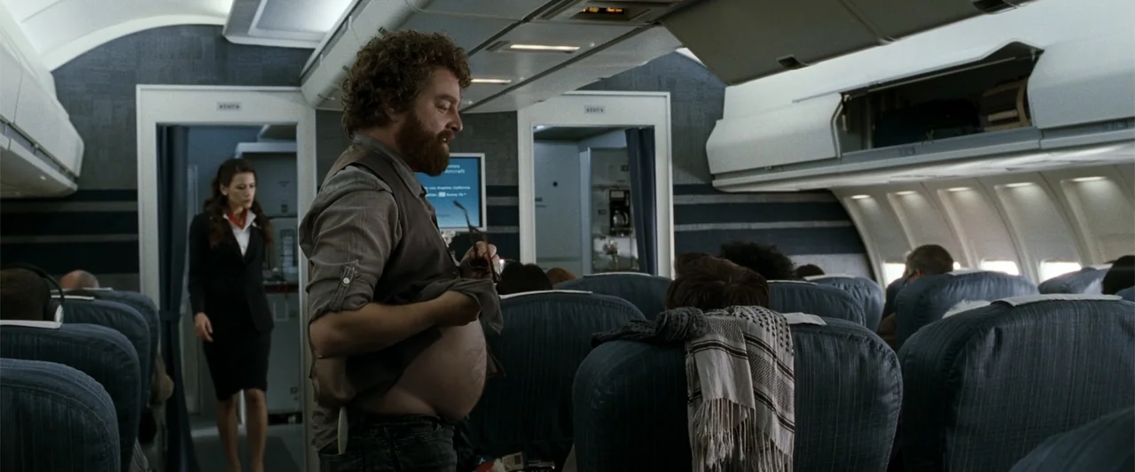 Due Date (2010) movie still: medium — The interior of a commercial airplane cabin features rows of blue seats and overhead bins. A man stands in the…