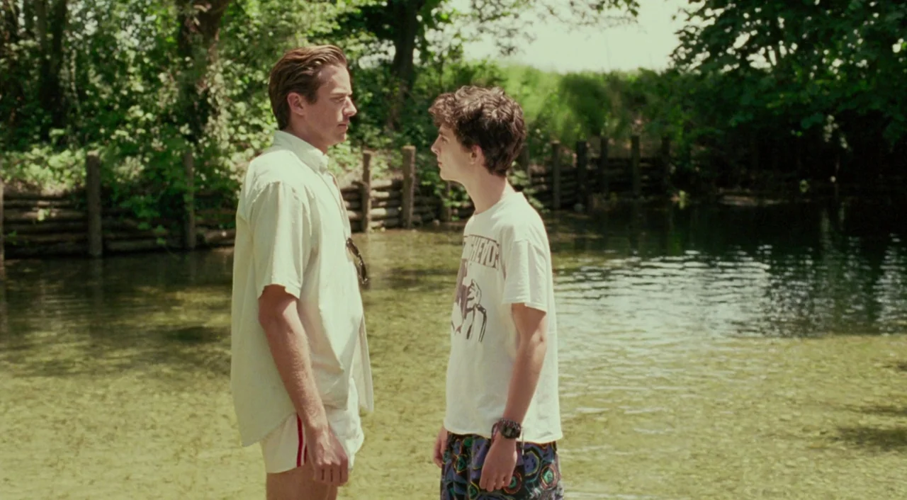 Call Me By Your Name (2017) movie still: fullbody — A shallow river with clear water runs through a lush green wooded area. Two young men stand in…