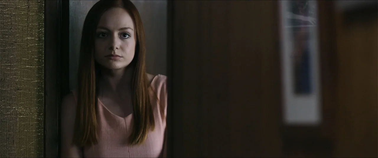 Captive (2020) movie still: medium — A dimly lit interior space features a wooden door frame partially obscuring the view. A woman with long auburn…