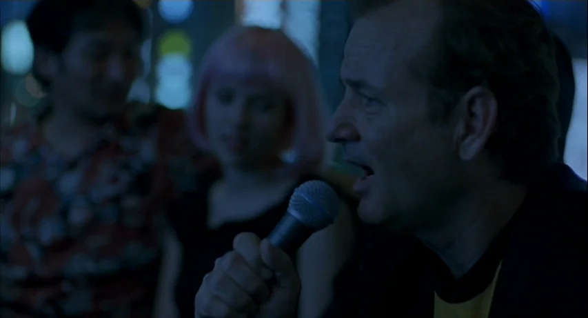 Lost In Translation (2003) movie still: medium — A dimly lit room with out-of-focus figures in the background. A man holds a microphone and sings…