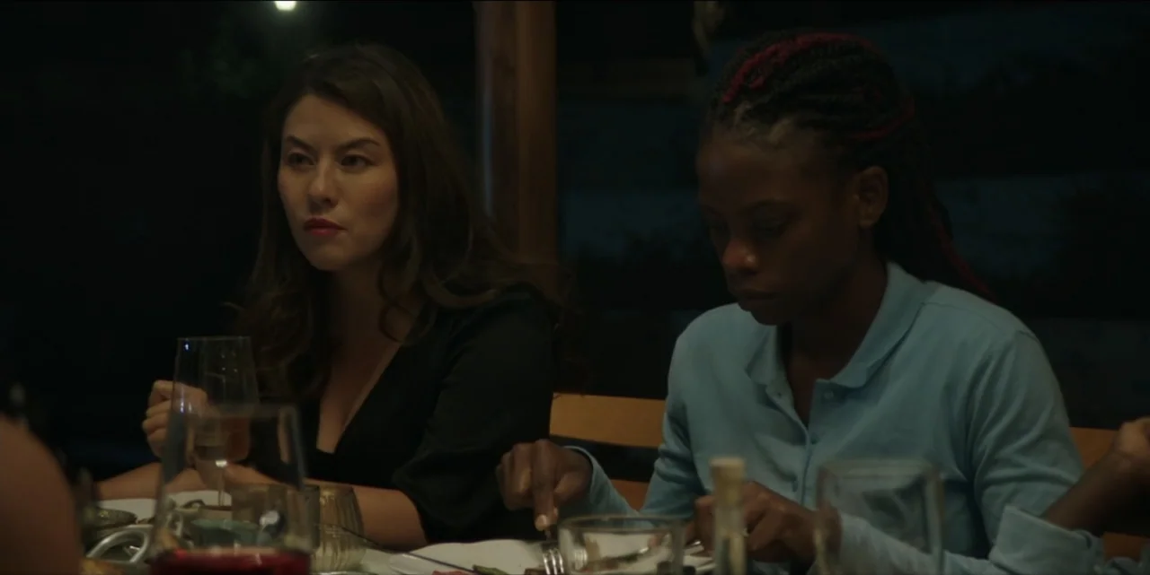 The Hunted (2024) movie still: medium — A dark dining room with a table set for a meal at night. Two women sit across from each other eating while…