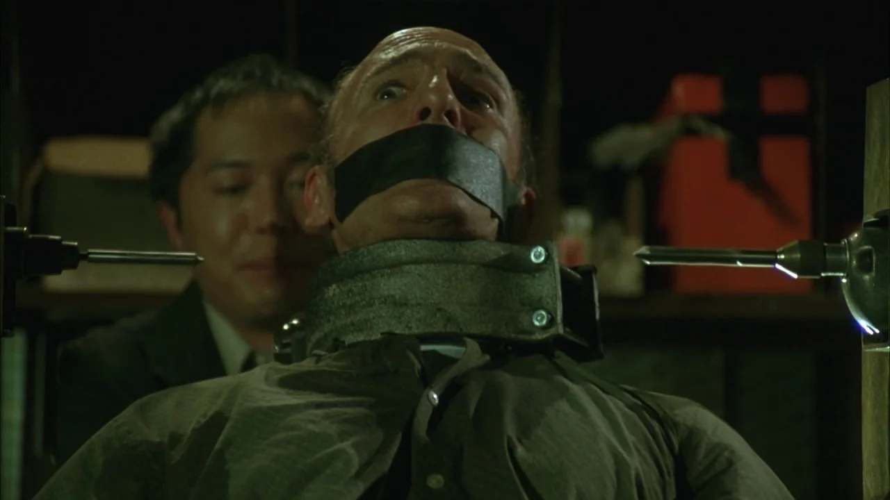 Saw movie still: closeup — A dimly lit room contains two men positioned near heavy machinery and power tools. One man wears a metal neck brace and…