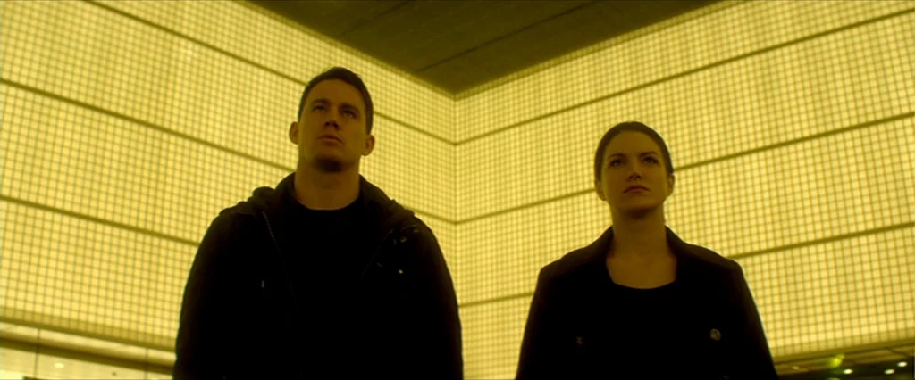 Haywire (2011) movie still: medium — An interior space features large yellow-lit modular wall panels forming a corner backdrop. A man and a woman…