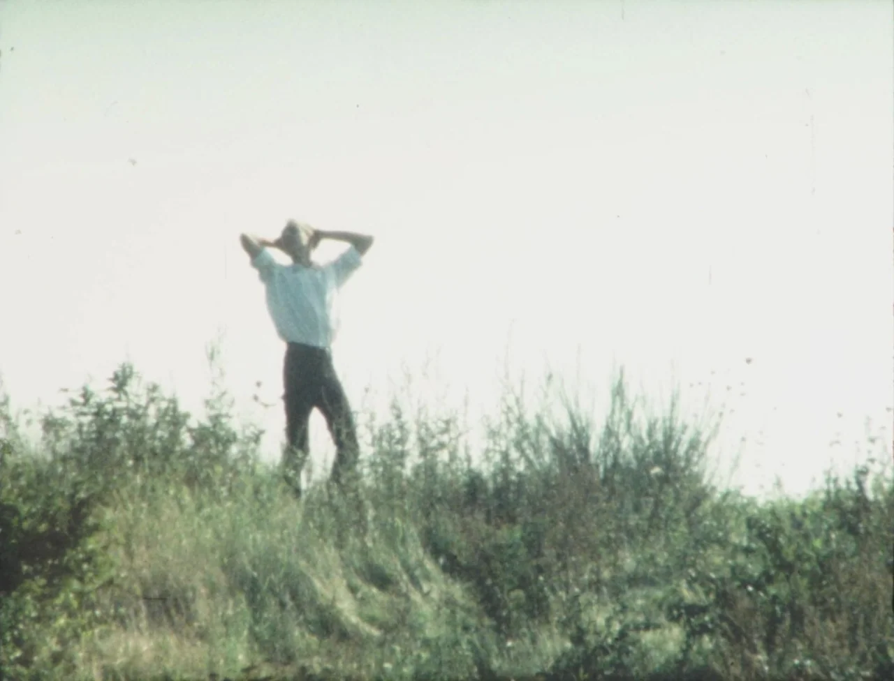 Nekromantik (1988) movie still: fullbody — A person stands in a grassy field against an overexposed, white sky. The subject poses with their hands…