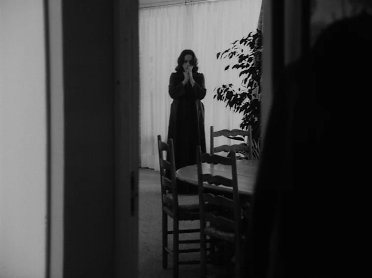 From the Life of the Marionettes (1980) movie still: medium — A dimly lit room contains a round wooden table surrounded by several chairs. A woman…