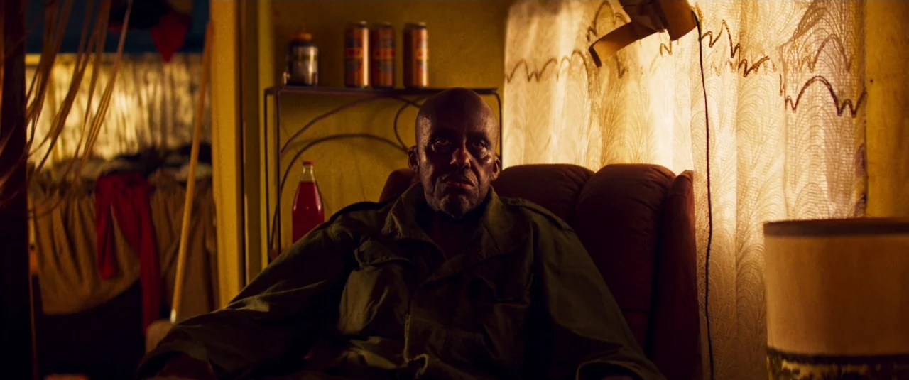 Mandy (2018) movie still: medium — A cramped interior with yellow walls and patterned curtains covering a window. An older man sits in a brown…