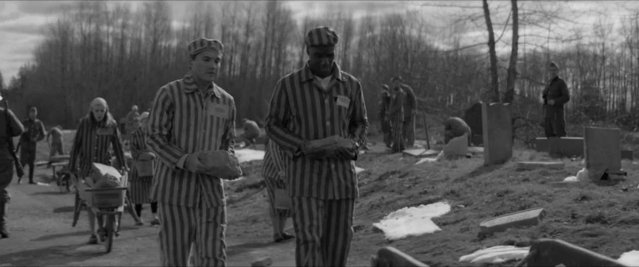 Bau, Artist at War (2025) movie still: fullbody — An outdoor area with bare trees, patches of snow, and multiple scattered gravestones. Two men…