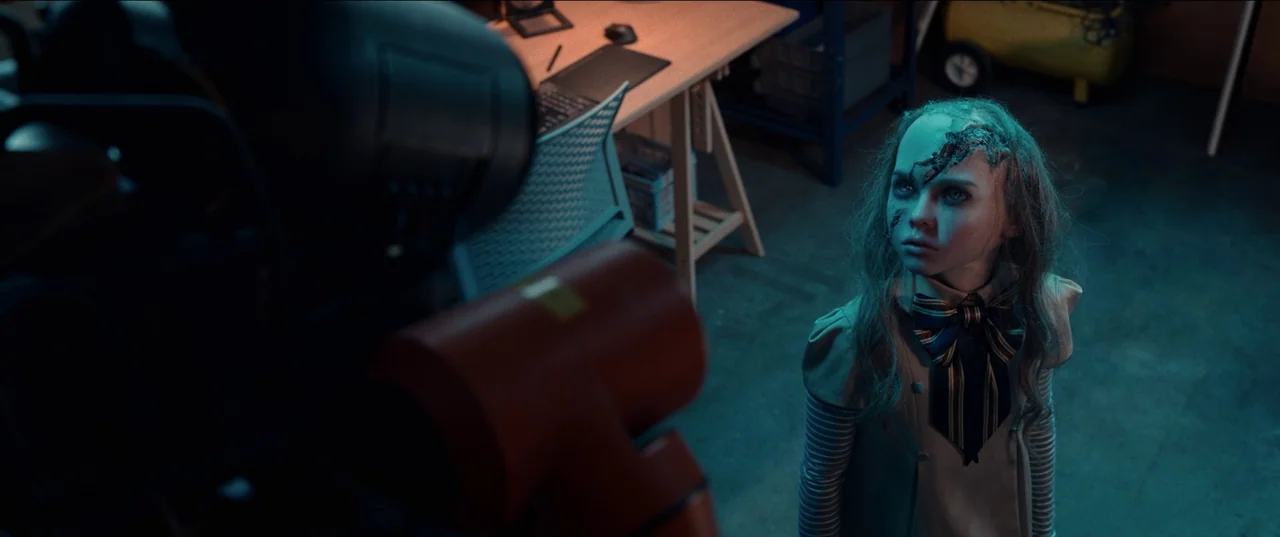 M3gan (2022) movie still: medium — A dimly lit workshop contains a mechanical arm towering over a damaged android doll. The android stands still…