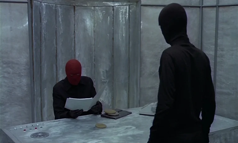 Nuits Rouges (1974) movie still: medium — A metallic, windowless room with curved walls and a smooth, reflective desk. A masked figure reviews…