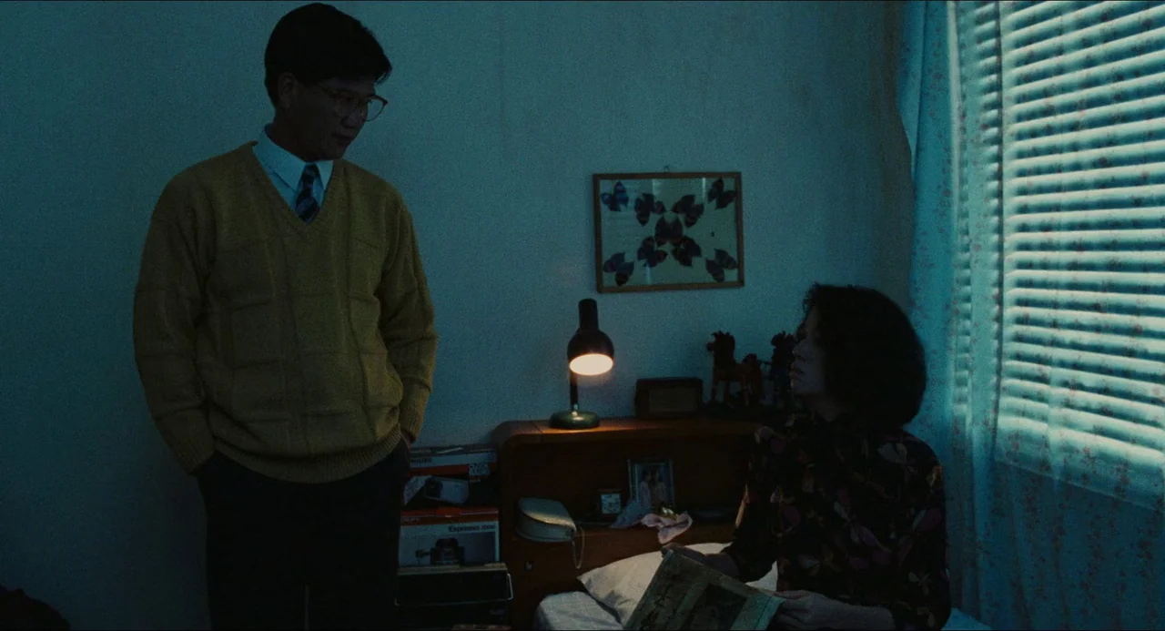 Rouge (1987) movie still: medium — A dimly lit bedroom features floral curtains over a window and a small desk lamp. A man stands over a seated…