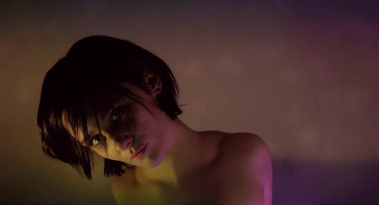 Like Me movie still: closeup — An interior space bathed in artificial multicolored lighting against a dark wall. A young woman with short dark hair…