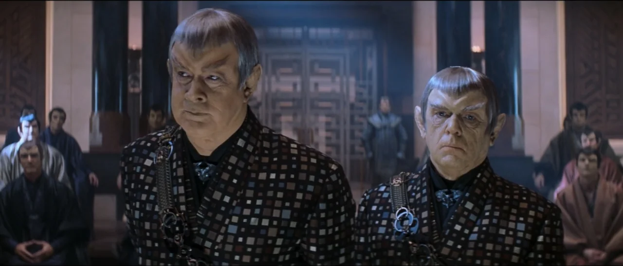 Star Trek: Nemesis (2002) movie still: medium — A formal council chamber features dark pillars, stone walls, and large metallic lattice doors. Two…