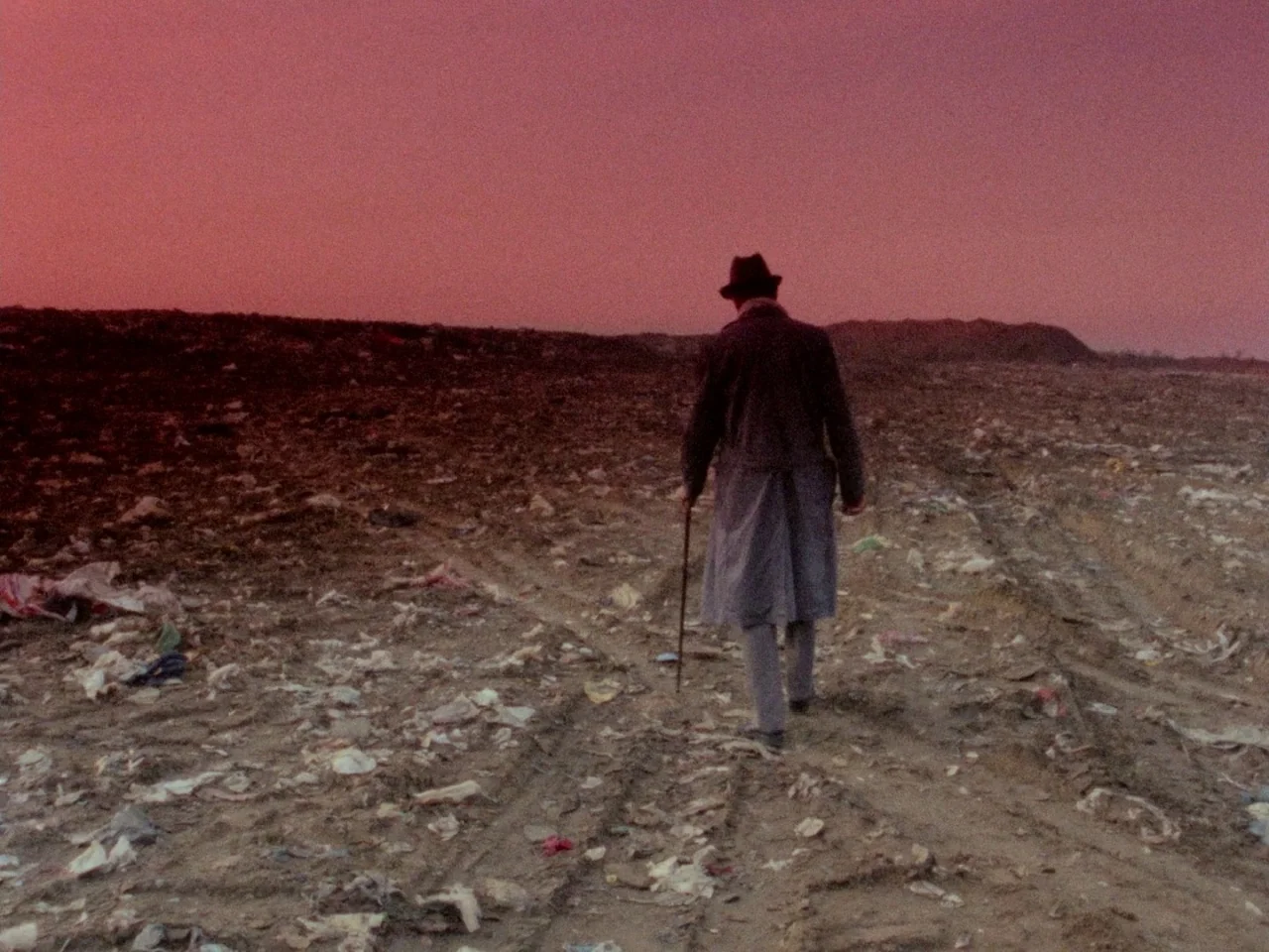 Decoder (1984) movie still: fullbody — A vast, barren landfill covered in scattered refuse sits beneath a deep reddish sky. A lone man wearing an…