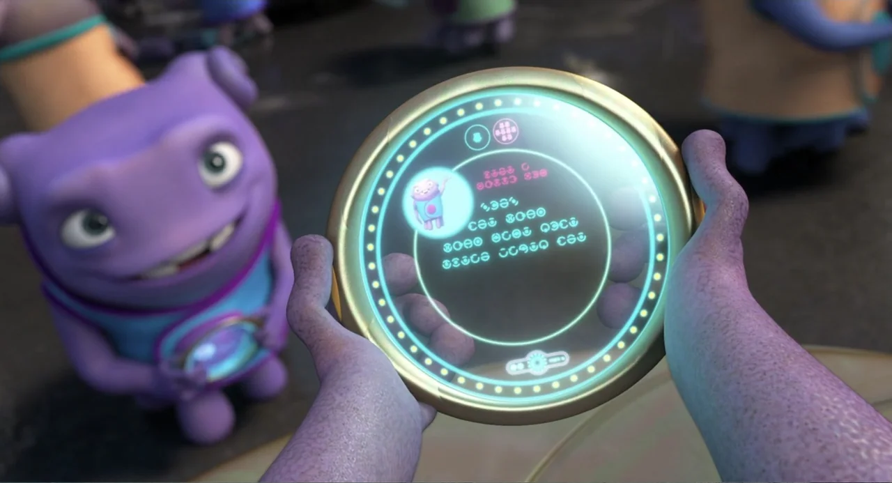 Home (2015) movie still: closeup — A textured purple hand holds a glowing circular device featuring alien symbols and text. In the background, a…