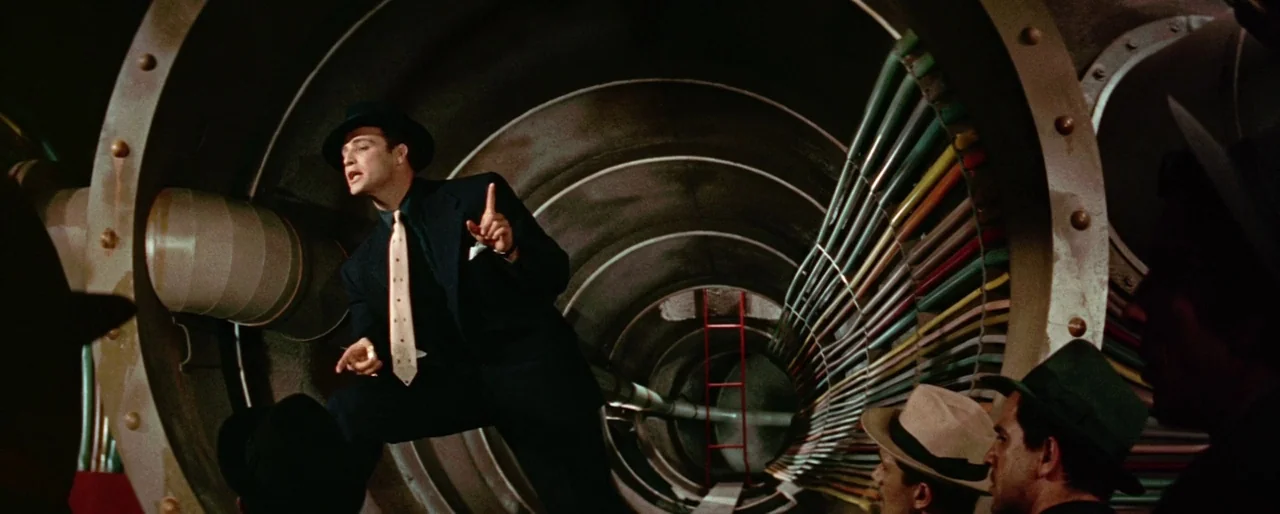 Guys and Dolls (1955) movie still: medium — A man stands before a large, circular, metallic industrial turbine or ventilation pipe. He addresses a…
