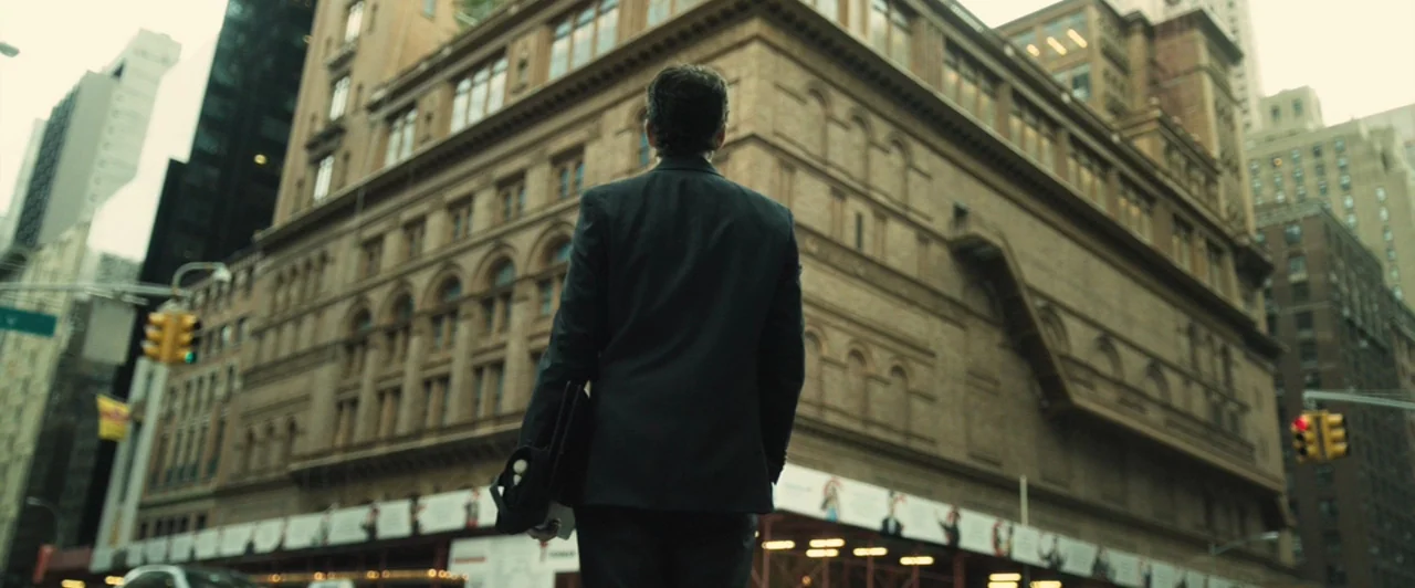 Whiplash (2014) movie still: fullbody — A man stands on a busy New York city sidewalk facing a massive brick building. He looks upward at the ornate…