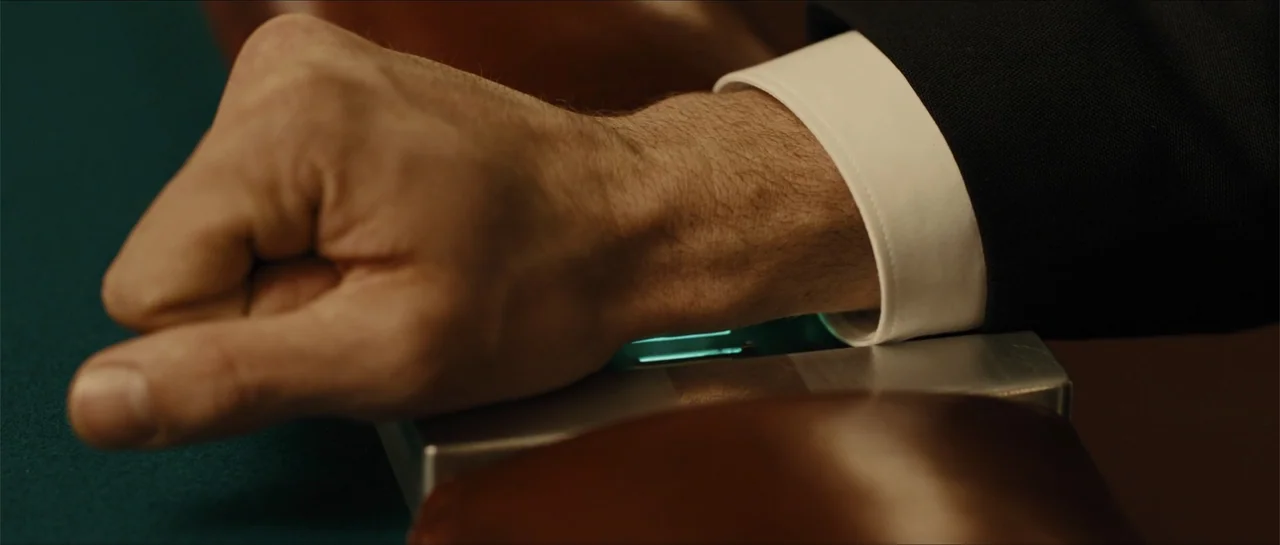 In Time (2011) movie still: closeup — A close view of a hand resting on a metal device atop a green felt table. The hand remains still on the…