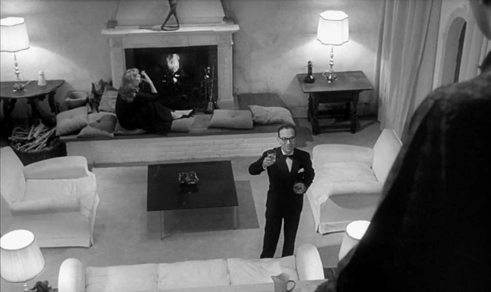 Eva (1962) movie still: wide — A spacious room features a stone fireplace, white armchairs, and a low central table. A man in a tuxedo stands…