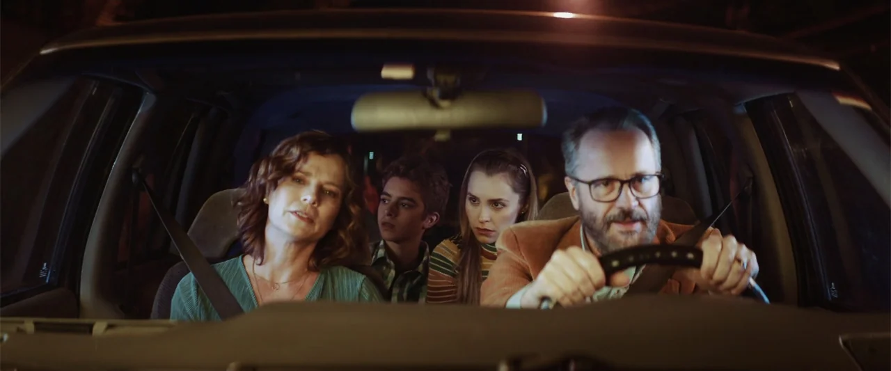 The Boy Who Killed My Parents (2021) movie still: medium — The interior of a moving car at night contains a family of four passengers. A man drives…