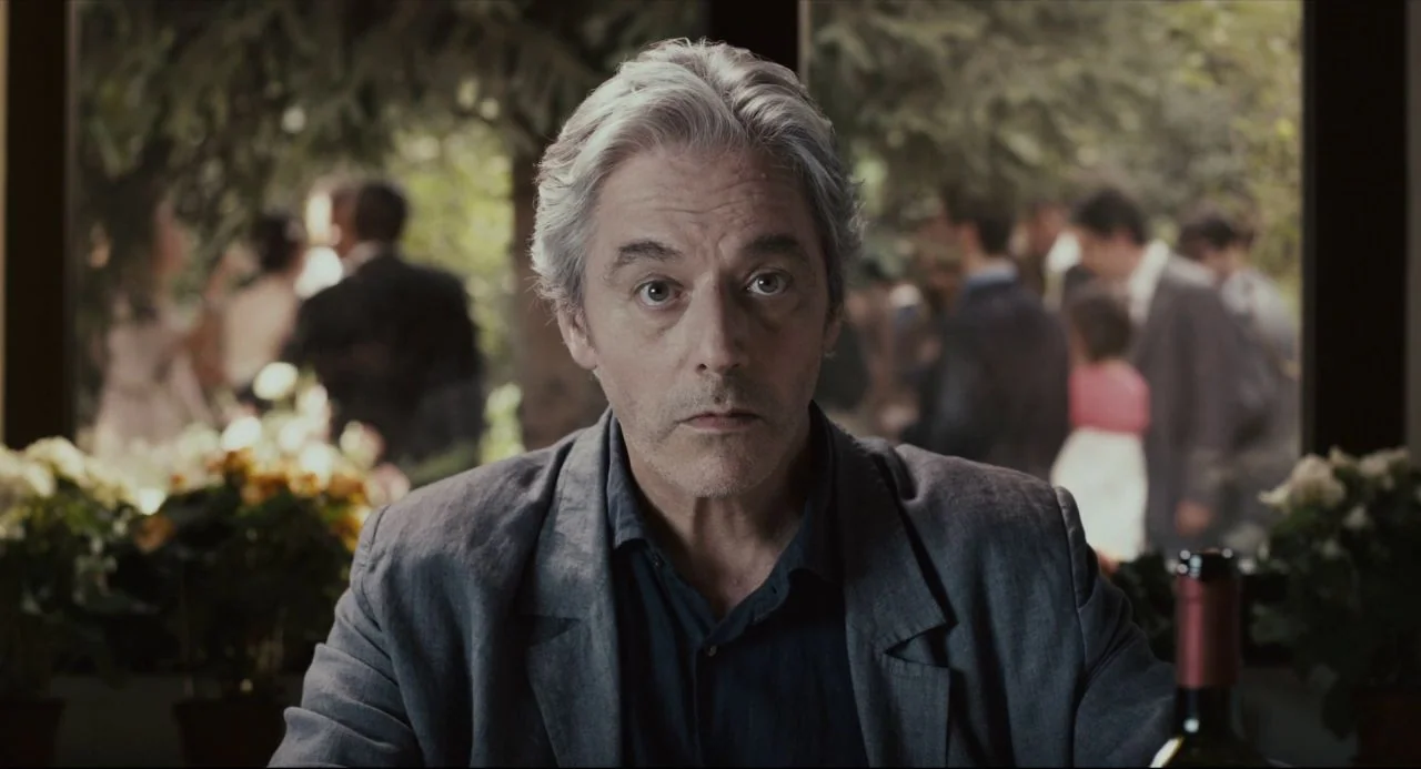 Certified Copy (2010) movie still: medium — A man sits at a table inside a space with large windows overlooking trees. He is positioned in the…