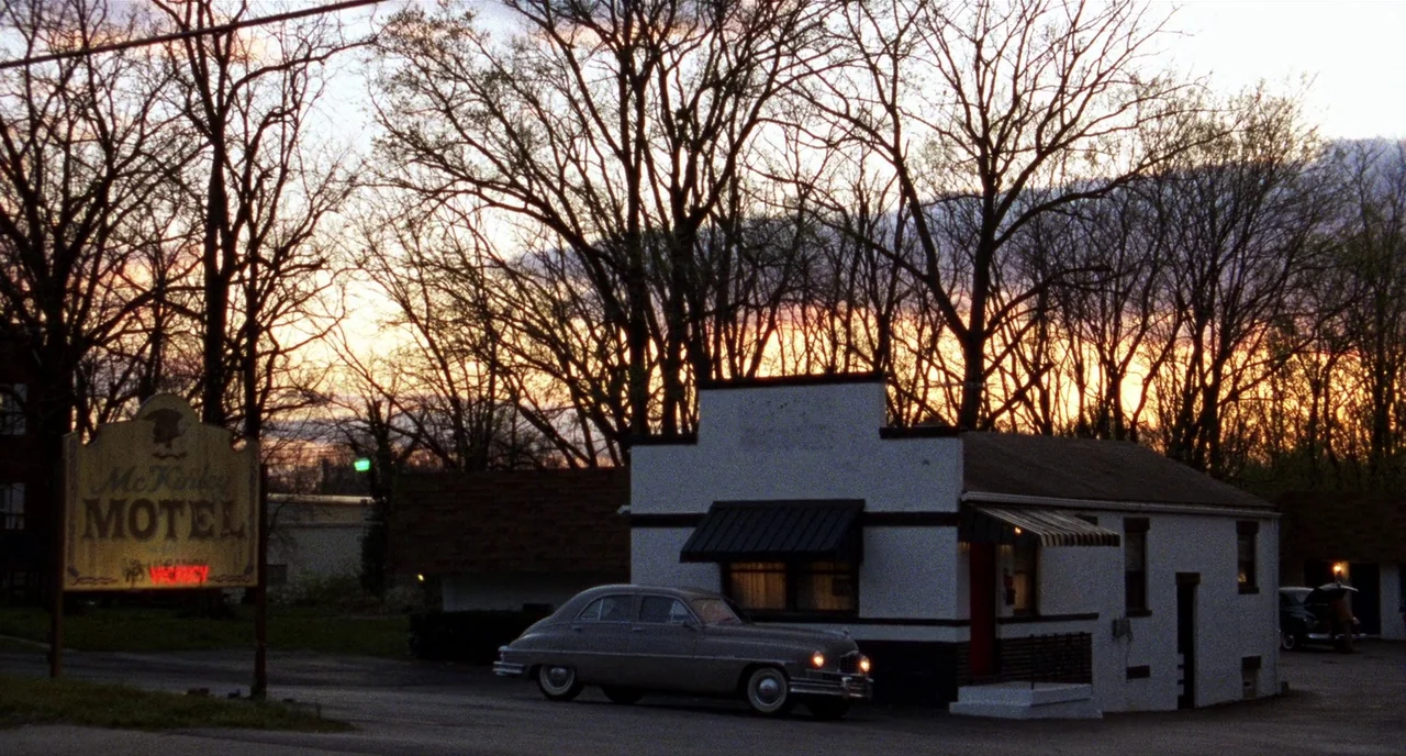 Carol (2015) movie still: wide — A small white motel building stands before a dense backdrop of leafless silhouetted trees. A vintage car is parked…