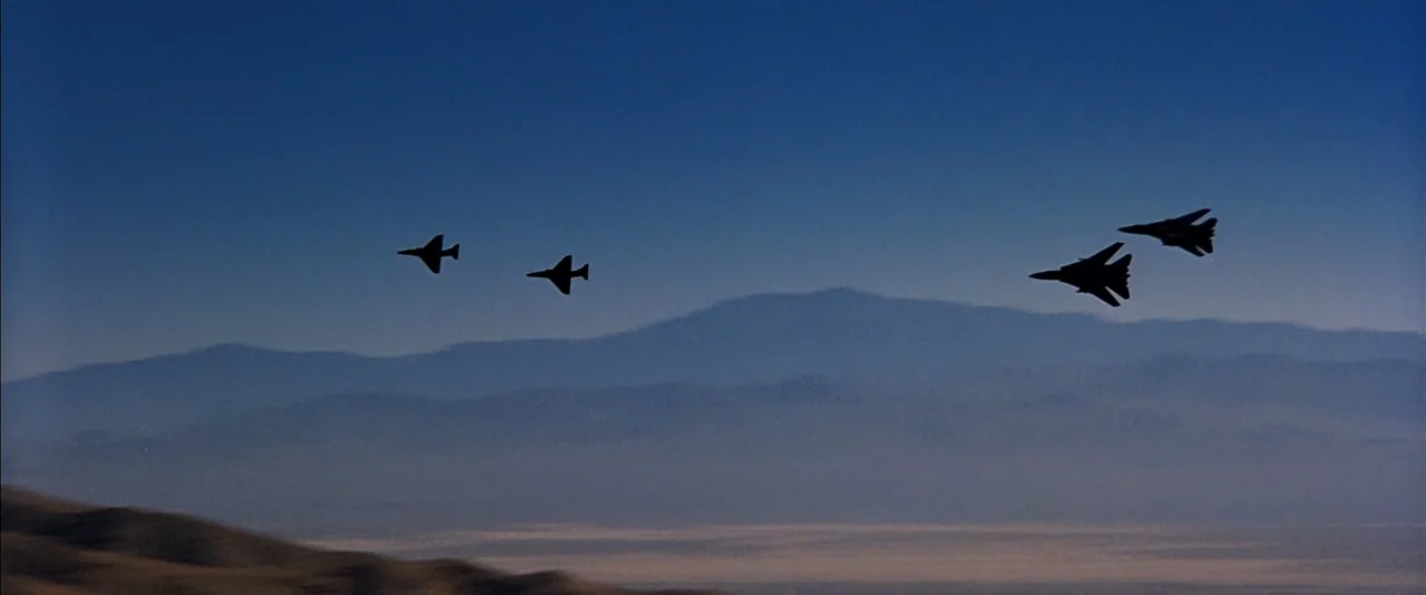 Top Gun (1986) movie still: wide — Four silhouetted fighter jets fly in formation against a clear, deep blue sky. Below the jets, a hazy desert…