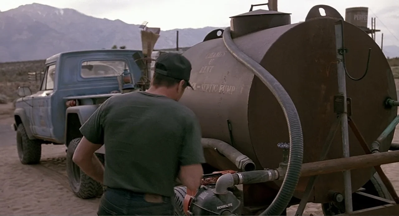 Tremors (1990) movie still: medium — A dry, dusty desert environment features a blue pickup truck and a septic tank. A man in a dark green t-shirt…