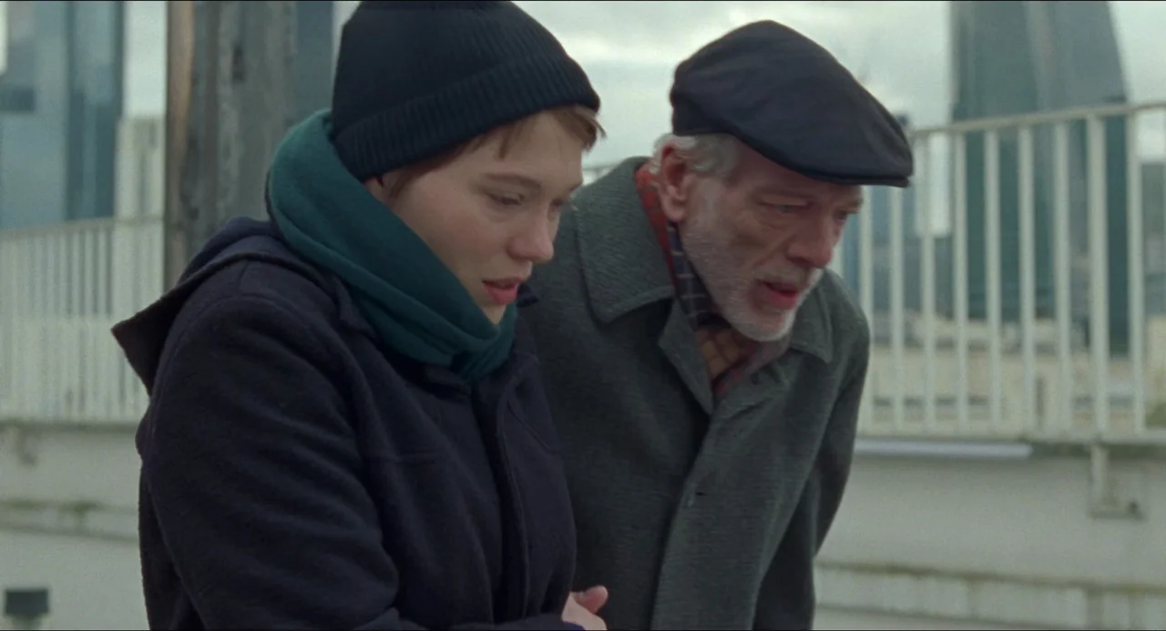 One Fine Morning (2022) movie still: medium — A woman and an elderly man stand together on a concrete pedestrian bridge. Both look down while city…