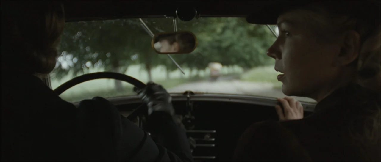 Suite Française (2015) movie still: medium — Two individuals sit inside a vintage automobile traveling along a tree-lined road. The passenger turns…