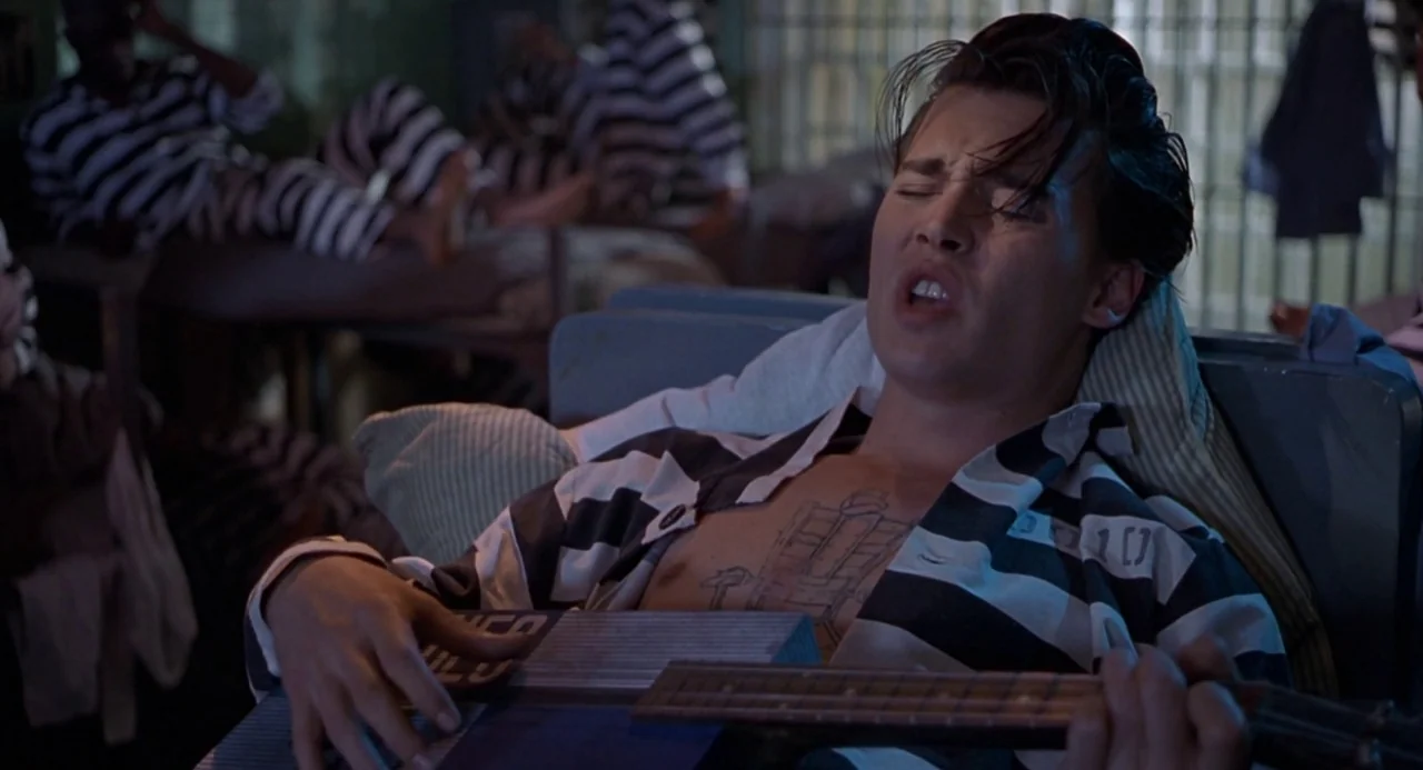Cry Baby (1990) movie still: medium — A dark jail cell contains multiple bunk beds with other inmates resting in the background. A young man sits on…