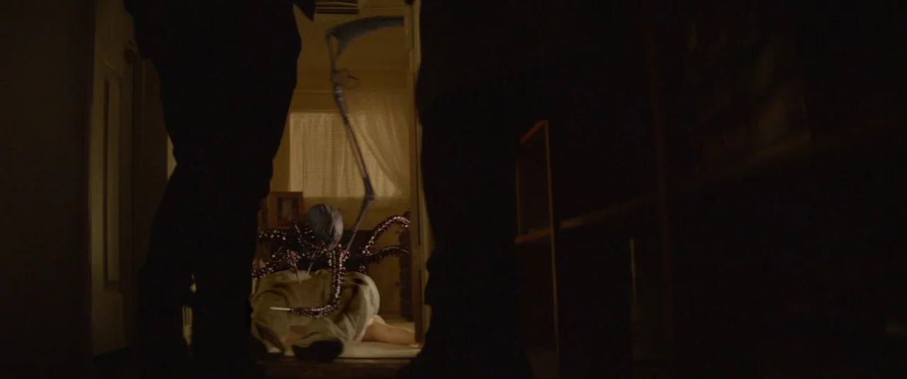 Southbound (2015) movie still: medium — A dimly lit narrow corridor leads to a room where a person lies prone. A multi-tentacled, dark creature…