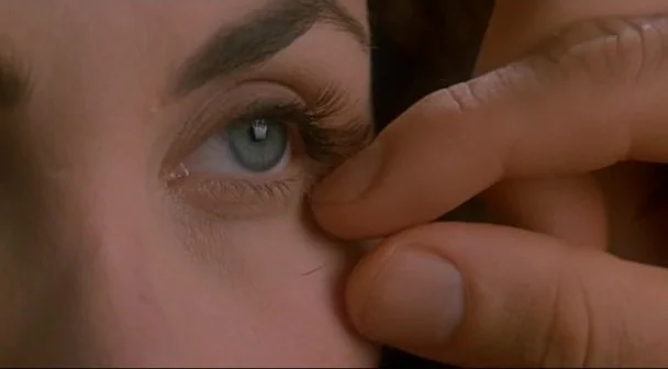 Boxing Helena (1993) movie still: closeup — A close-up view shows a person's finger gently touching the skin under another's eye. The eye with blue…