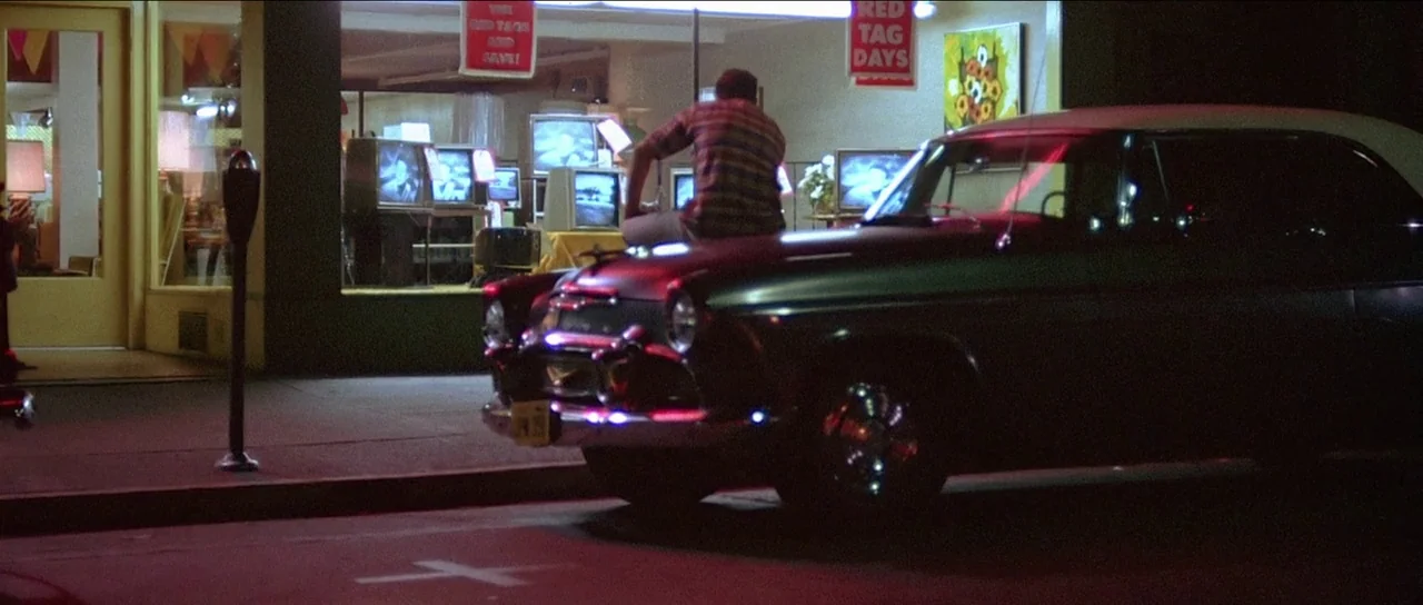 American Graffiti (1973) movie still: wide — A night scene outside a retail store illuminated by interior display lights and signs. A man sits alone…