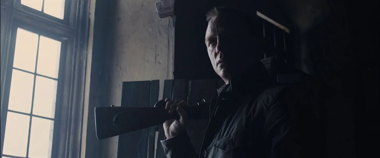 Skyfall (2012) movie still: medium — A man stands indoors next to a large window in a darkened, rustic building. He holds a long rifle over his…