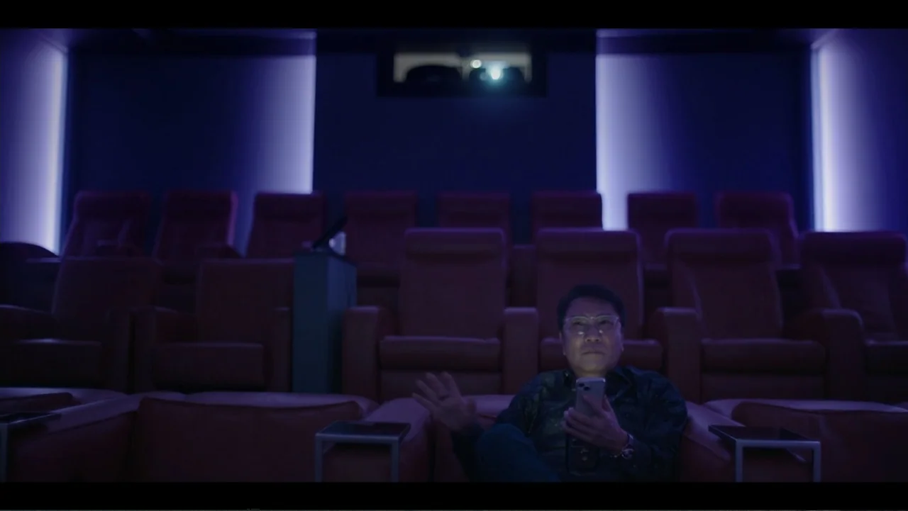 Lee Soo Man: King of K-Pop (2025) movie still: medium — A dimly lit modern movie theater with rows of empty plush red seats. A man sits alone in a…
