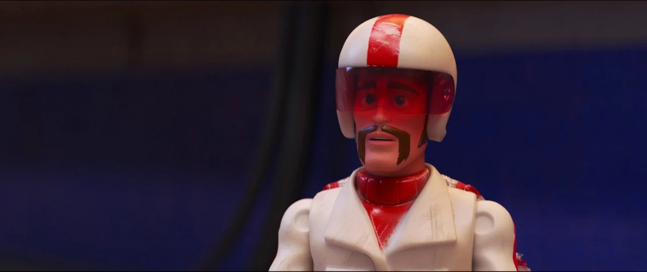 Toy Story 4 (2019) movie still: medium — A plastic action figure stands against a plain blue background surface. The figure wears a white racing…