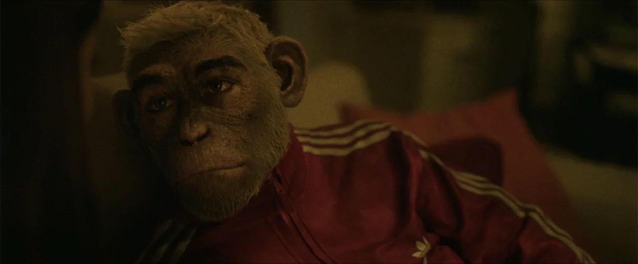 Better Man (2024) movie still: closeup — A dimly lit room interior contains a seated humanoid chimpanzee. The figure wears a red athletic jacket…