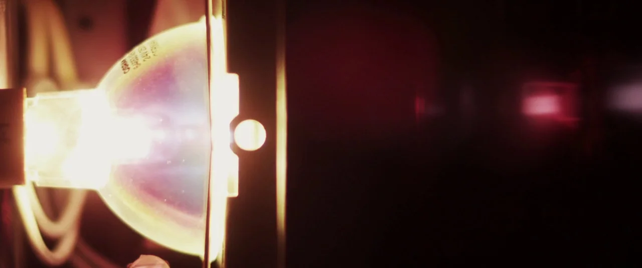 The Canal (2014) movie still: closeup — A close view of a glowing halogen bulb encased in a metal fixture. The light source emits a bright flare…