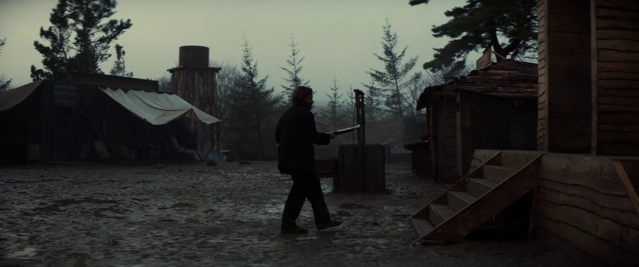 Never Grow Old (2019) movie still: wide — A muddy, underdeveloped frontier encampment with primitive wooden structures and canvas-covered shelters.…