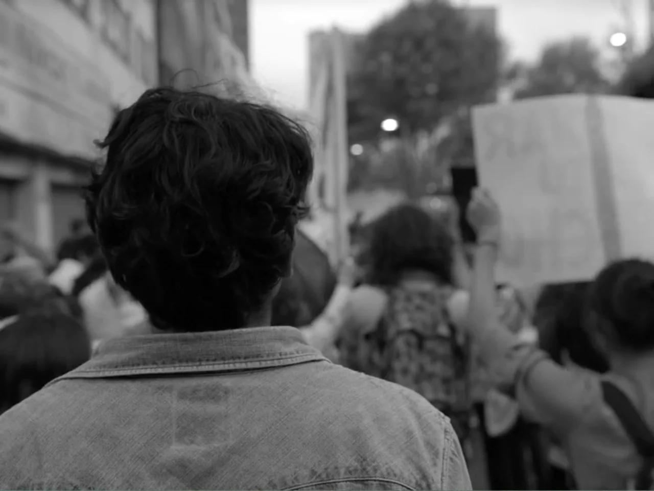 Güeros (2014) movie still: medium — A city street packed with people participating in a public protest at dusk. A person in a denim jacket stands in…