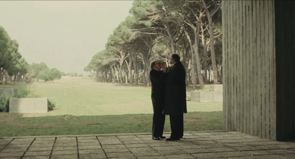 Todo Modo (1976) movie still: fullbody — A stone-paved patio borders a wide, grassy field lined with tall, slender pine trees. Two men in dark…