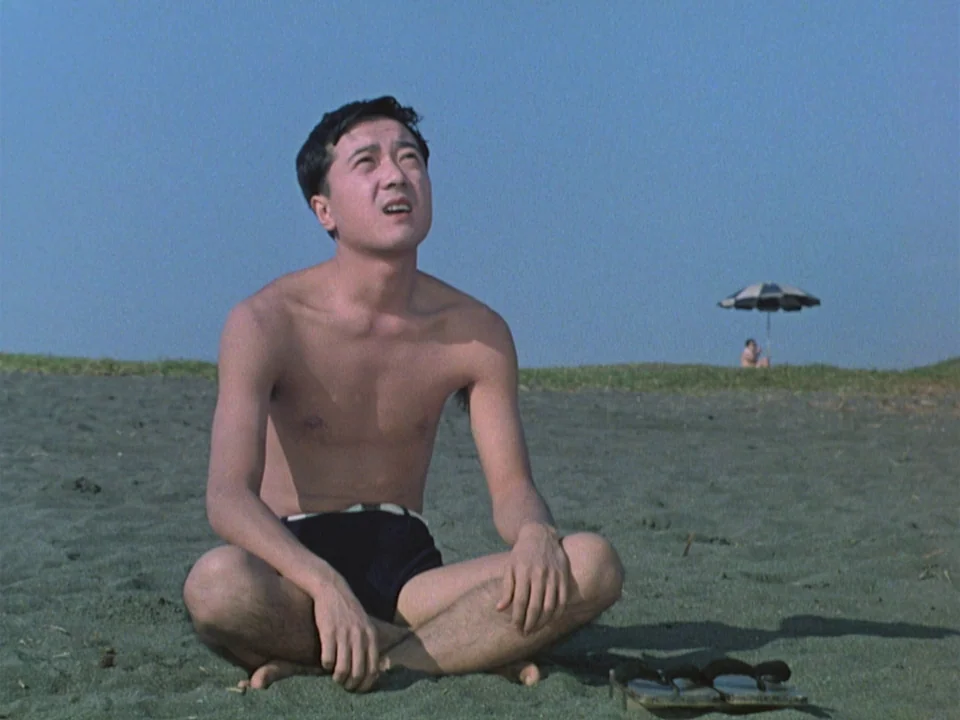 Floating Weeds (1959) movie still: medium — A flat stretch of dark sand sits under a clear blue sky. A young man sits cross-legged while looking…